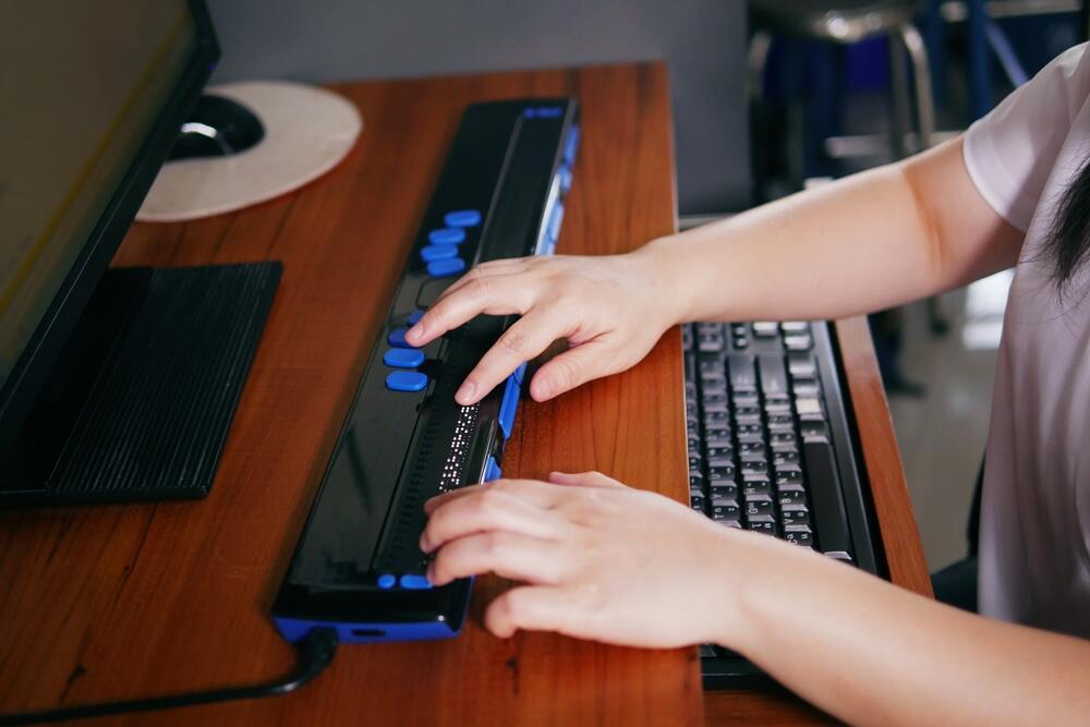A person with blindness uses a computer via a Braille assistive device, which can be used with screen reader software to browse websites.