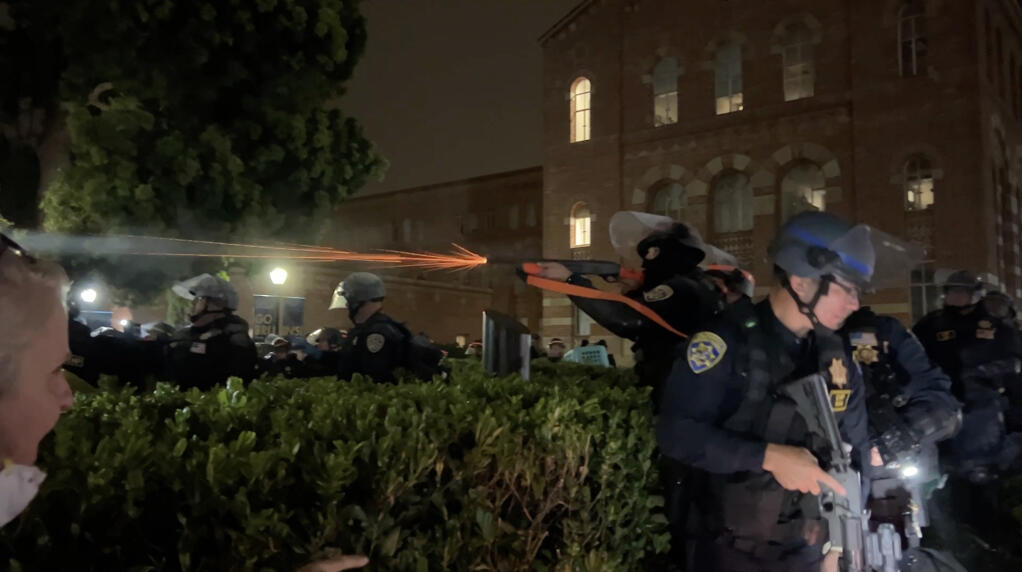 An officer fires “impact munitions” into a crowd of pro-Palestinian protesters at UCLA during the early morning of May 2, 2024, as police cleared an encampment that protesters had refused to vacate. Screenshot via a video by Sergio Olmos, CalMatters