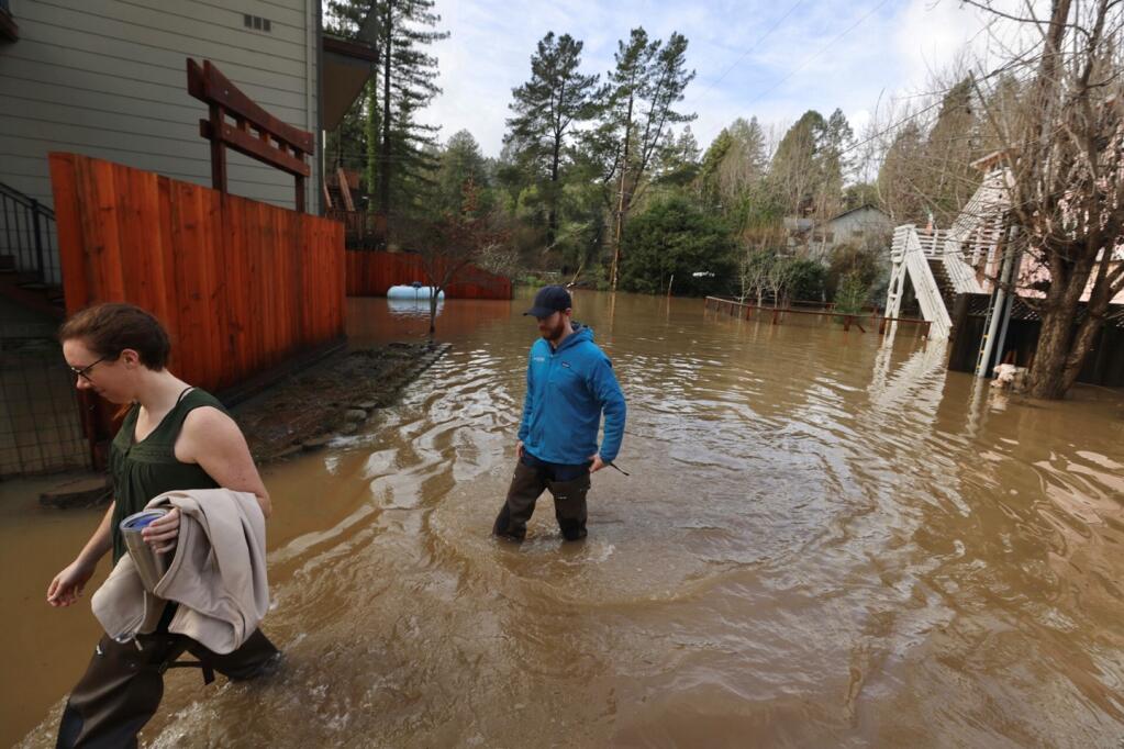 Meg Cooper and George Muldoon walk through floodwaters from Fife Creek to get to their home on Mill Court in Healdsburg, Monday, Jan. 9, 2023. (Beth Schlanker/The Press Democrat)