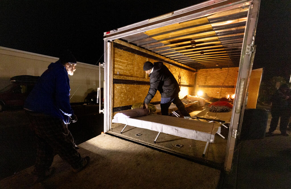 Richard Jump holds a light for Ray Joseph as he sets up cots in a retired Need a Hand Movers moving van now repurposed as a warming station for homeless people on Santa Rosa  and East Robles avenues in Santa Rosa, Tuesday, Feb. 6, 2024. (John Burgess / The Press Democrat)
