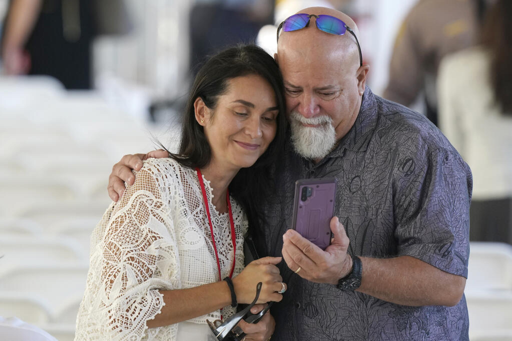 Two attendees gather around a phone before the start of a remembrance event at the site of the Champlain Towers South building collapse, Friday, June 24, 2022, in Surfside, Fla. Friday marks the anniversary of the oceanfront condo building collapse that killed 98 people in Surfside, Florida. The 12-story tower came down with a thunderous roar and left a giant pile of rubble in one of the deadliest collapses in U.S. history. (AP Photo/Wilfredo Lee)