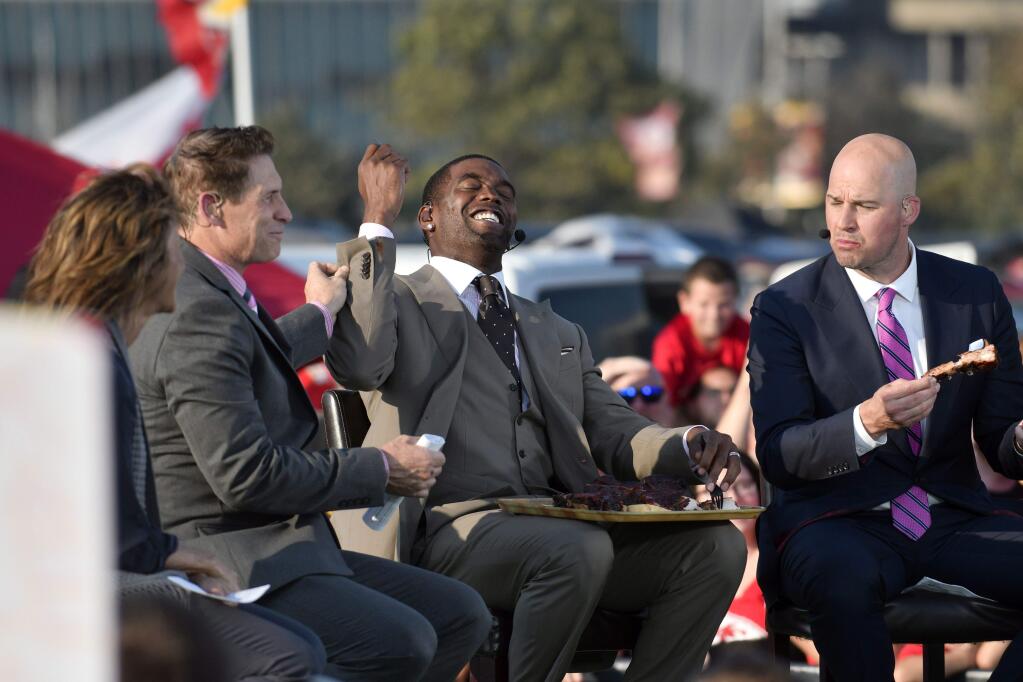ESPN host Steve Young, left, holds the sleeve of Randy Moss, center, who has bbq sauce on it, as Matt Hasselbeck, right, samples food during a tailgating special outside Arrowhead Stadium before an NFL football game between the Kansas City Chiefs and the Washington Redskins, in Kansas City, Mo., Monday, Oct. 2, 2017. (AP Photo/Ed Zurga)