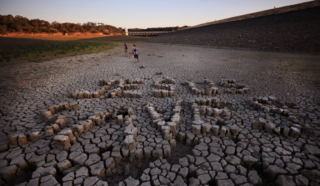 At the foot of Coyote Valley Dam on the bottom of Lake Mendocino, someone spelled out “Jesus Saves” with drought-cracked blocks of mud, Thursday, July 15, 2021 east of Ukiah. In the background are Ukiah residents Hannah Campion, Aven Bevilacqua and Jason Griego with their dog Apollo. (Kent Porter / The Press Democrat) 2021