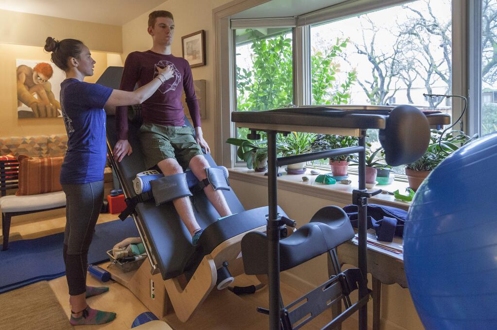 Theo St. Francis get some assistance from physical therapist Kinley McCracken on a piece of specialized equipment that helps with the motion of walking. The family living room was transformed into a fitness room with specialized equipment. (Photos by Robbi Pengelly/Index-Tribune)