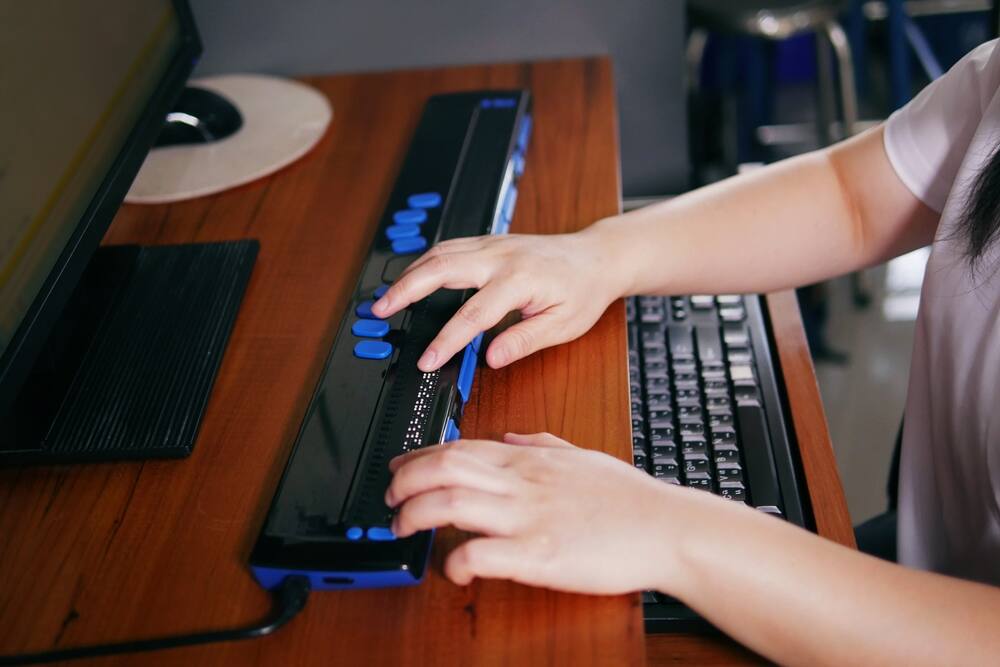 A person with blindness uses a computer via a Braille assistive device, which can be used with screen reader software to browse websites.