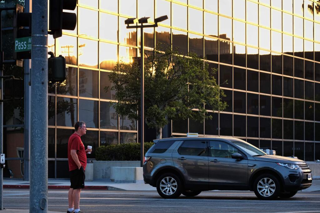 In this Sept. 21, 2018 photo a pedestrian with his morning cup of coffee in hand crosses a street as the sun rises in the reflection of a building in Burbank, Calif. It's clockwork: Every fall and spring, Californians and the rest of the nation switch to daylight saving or standard time, often with gripes about losing an hour of sleep, the sun setting earlier in the day or the chore of changing analogue clocks. This November, California voters will get to decide if it's time for a change. If voters approve Proposition 7 on Nov. 6, that would pave the way for year-round daylight saving time in the state. (AP Photo/Richard Vogel)