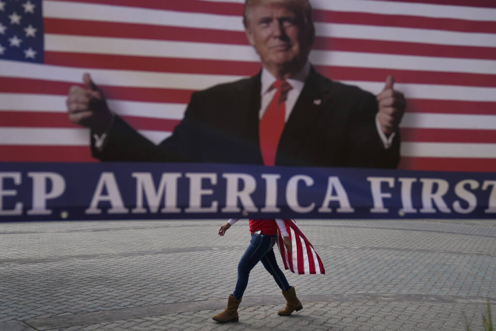 A woman draped in an American flag walks past a banner supporting President Donald Trump during a rally Wednesday, Jan. 6, 2021, in Huntington Beach, Calif. (AP Photo/Jae C. Hong)