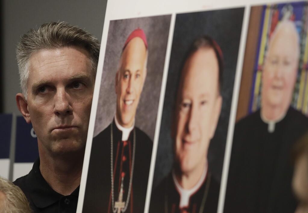 Tom Emens listens to speakers as he stands next to photos of San Francisco Archbisop Salvatore Cordileone, from left, Oakland Bishop Michael Barber and San Jose Bishop Patrick McGrath at a news conference in San Francisco, Tuesday, Oct. 23, 2018. A law firm suing California bishops for the records of priests accused of sexual abuse has compiled a report of clergy in the San Francisco Bay Area it says are accused of misconduct. (AP Photo/Jeff Chiu)