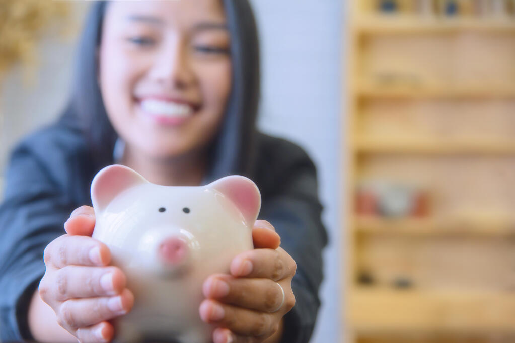 A young Asian woman holds a piggy bank in her hand hands with a wooden bookcase in the background.