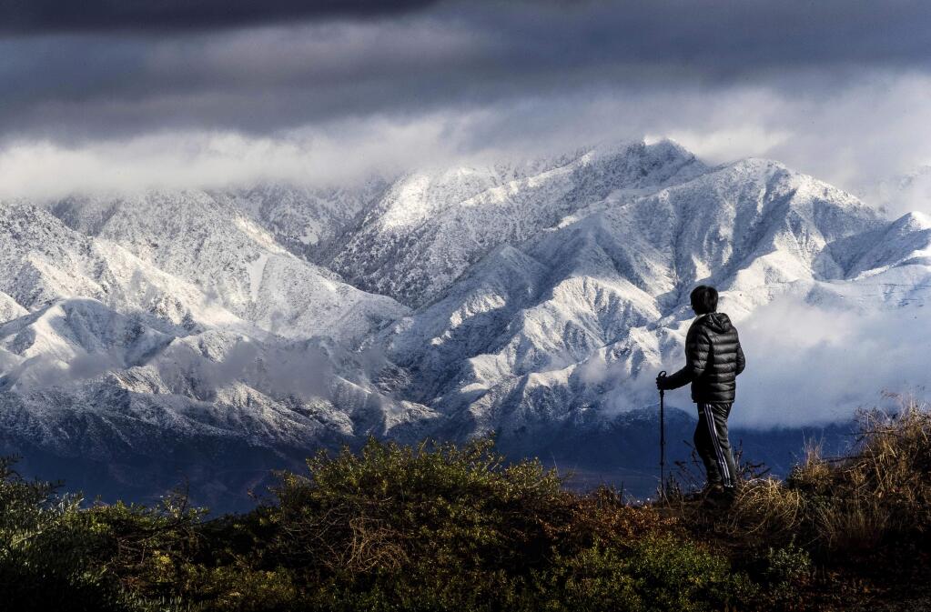 Frank Huong takes a break from hiking to enjoy a view of snow-capped San Gabriel Mountains from Hidden Hills trail in Chino Hills, Calif., on Friday, Nov. 29, 2019. Rain and snow showers are continuing in parts of the state Friday morning while skies are clearing elsewhere. ( Watchara Phomicinda/The Orange County Register via AP)