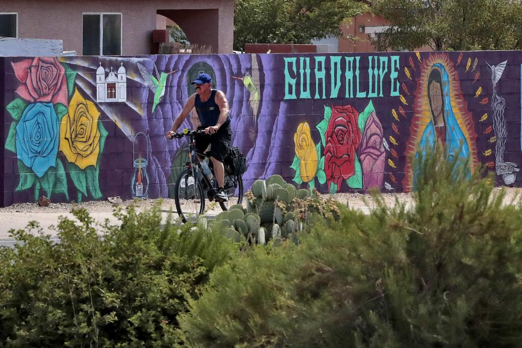 In this Saturday, June 13, 2020, photo, a man cycles past a mural in Guadalupe, Ariz. As the coronavirus spreads deeper across America, it's ravaging through the homes and communities of Latinos from the suburbs of the nation's capital to the farm fields of Florida to the sprawling suburbs of Phoenix and countless communities in between. (AP Photo/Matt York)