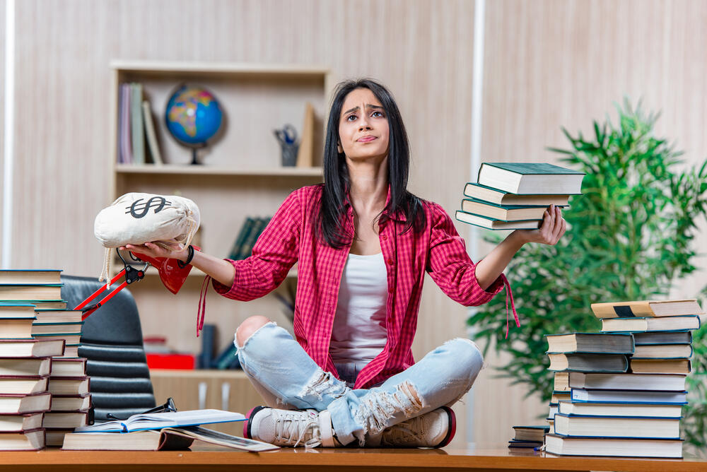 A young female student sits on a desk, surrounded by books and balancing a sack of money (financing or debt) in the right hand with college education (books) in the left hand.