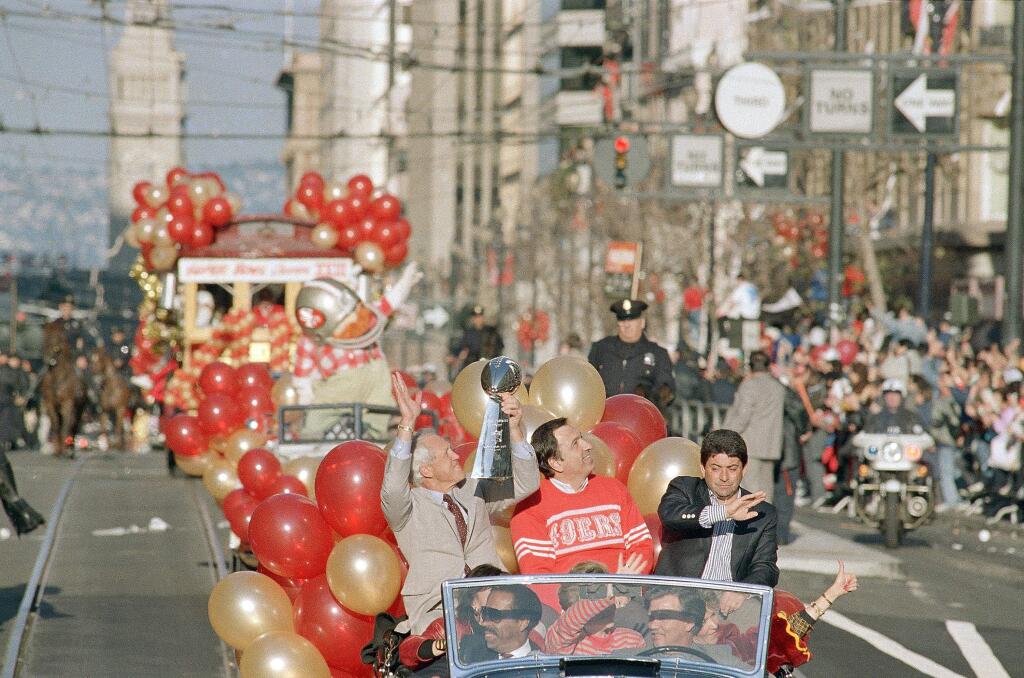 San Francisco 49ers coach Bill Walsh, left, holds the Super Bowl trophy aloft and waves to the crowd as he rides at the head of the welcome home parade with Mayor Art Agnos, center, and 49ers owner Eddie DeBartolo Jr., right, Jan. 23, 1989. Thousands lined Market Street for the victory parade which featured team members riding of motorized cable cars. (AP Photo/Paul Sakuma)
