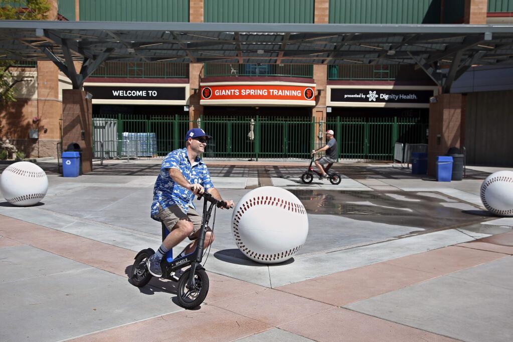 Joe Lieser, left, and Matt Kirmer, right, both of Torrance ride scooters past Scottsdale Stadium, the spring training home of the San Francisco Giants, in Scottsdale, Ariz., Saturday, March 14, 2020. The two came to Arizona with their families to attend spring training games but have not been able to attend any, as the remainder of the exhibitions have been canceled due to coronavirus pandemic. (AP Photo/Sue Ogrocki)