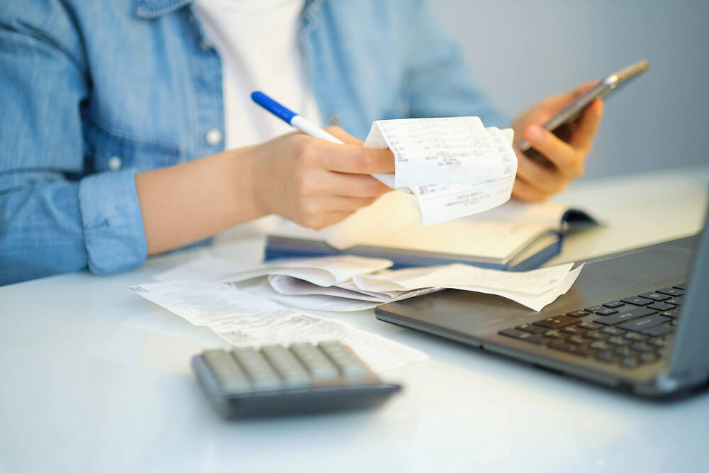 A woman, surrounded by a calculator, laptop and account book on a table, holds a pen, receipts and a smartphone.