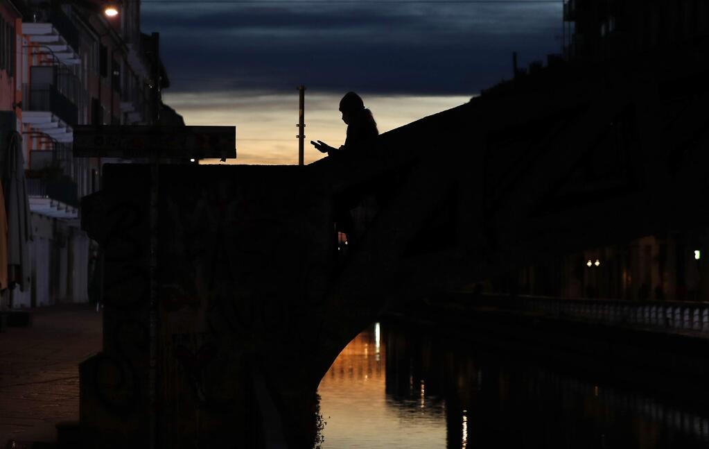 FILE -- In this March 10, 2020 file photo, a woman wearing a face mask checks her phone as she walks at the Naviglio Grande canal in Milan, Italy. Across Europe, governments are increasingly using surveillance to try to curtail the lethal spread of the new coronavirus, tracking people's movements with aggregated cell phone location data and introducing apps to keep the infected quarantined. (AP Photo/Antonio Calanni)