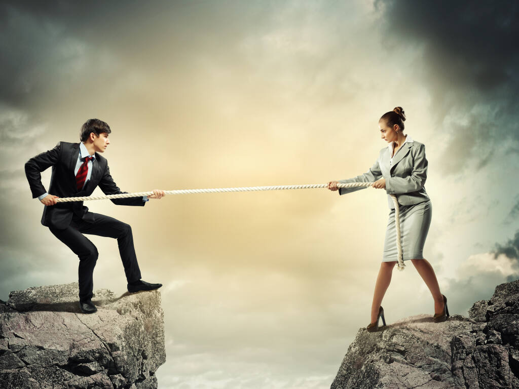 This collage image shows two white businesspeople in suits, a man on the left and a woman on the right, engage in tug of war with a rope across a gap between two rocky outcrops.