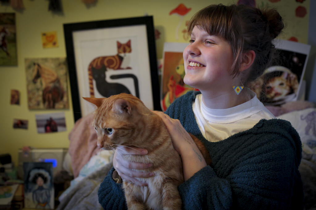 Aspiring artist, Vita Langst, a Petaluma High School sophomore, will be selling prints and stickers of her art at LumaCon this Saturday. Sitting in her bedroom with her cat, Henri, she is surrounded by some of her work that decorates her bedroom, including art that features Henri. _Tuesday, Jan. 24, 2023._(CRISSY PASCUAL/ARGUS-COURIER STAFF)
