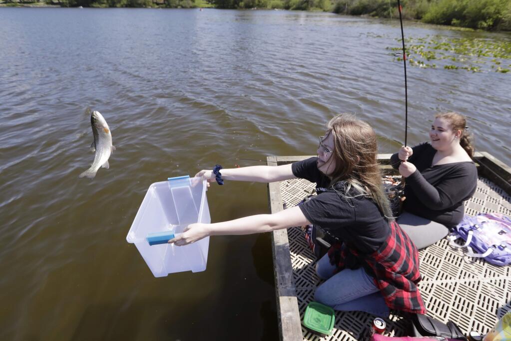 FILE - In this Tuesday, May 5, 2020, file photo, Anastasia Traicoff, left, reaches a bin toward a trout being reeled-in by her friend Julie Barnett as they fish in Blackmans Lake on the first day of a partial reopening of outdoor recreation activities in Snohomish, Wash. Fishing is seeing an increase in participation since the coronavirus outbreak began taking off in the United States (AP Photo/Elaine Thompson, File)