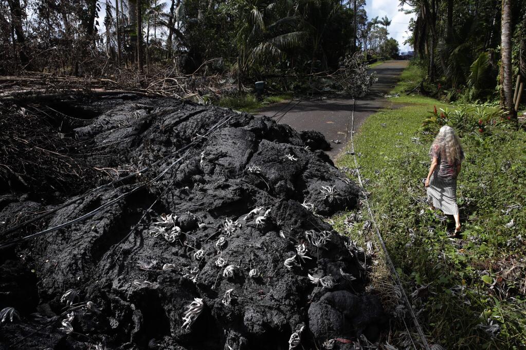 FILE- In this May 11, 2018 file photo, Hannique Ruder, a 65-year-old resident living in the Leilani Estates subdivision, walks past the mound of hardened lava while surveying the neighborhood near Pahoa, Hawaii. As Hawaii's Kilauea volcano sputters lava into a second week, scientists are examining samples to seek clues on what might be happening beneath the earth's surface. The age of the lava and the rate at which it's shooting out of the ground also provides hints on the length and intensity of the current eruption. Sometimes the spurts of lava are shooting fountains 200 feet into the air. (AP Photo/Jae C. Hong, File)
