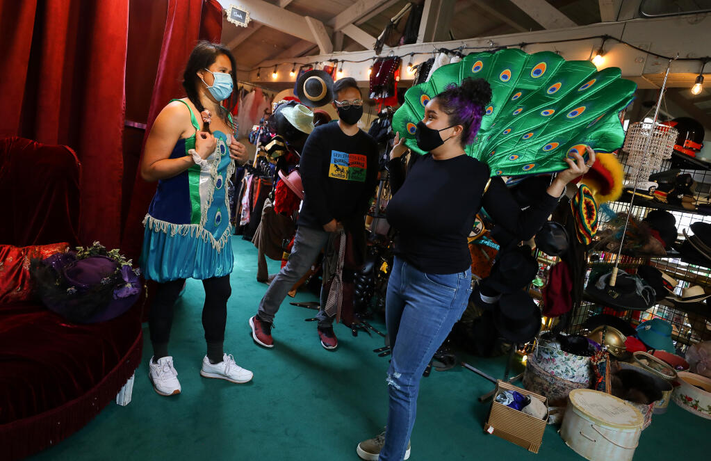 Store manager Iliana Sanchez, right, shows Andrea De Leon an optional way to wear an accessory on a peacock costume, while Eddie De Leon watches, at Disguise the Limit in Santa Rosa on Friday, April 23, 2021.  The owner of the building is selling the property, and the costume shop must find a new location.  (Christopher Chung/ The Press Democrat)