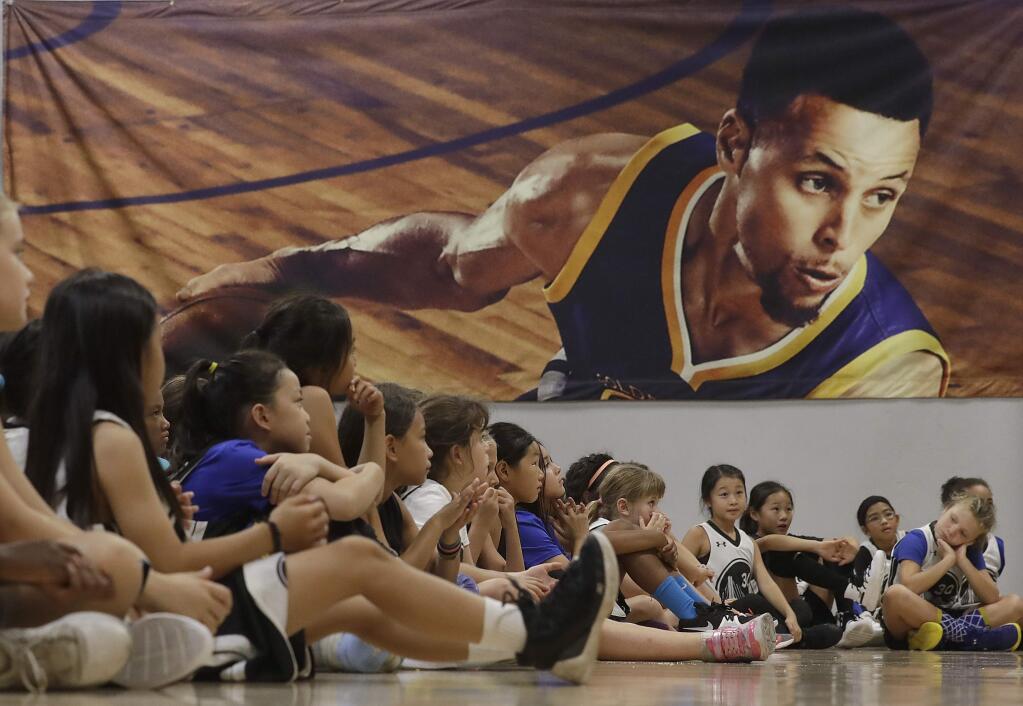 An image of Golden State Warriors' Stephen Curry, top, is displayed behind basketball camp participants as they listen to speakers on a panel for women in sports at Ultimate Fieldhouse in Walnut Creek, Tuesday, Aug. 14, 2018. (AP Photo/Jeff Chiu)