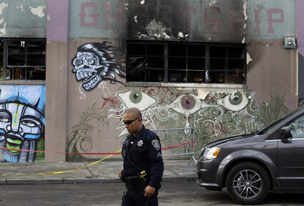 FILE - In this Dec. 9, 2016 file photo, an Oakland police officer guards the area in front of the art collective warehouse known as the Ghost Ship in the aftermath of a fire in Oakland, Calif. Max Harris and Derick Almena, the two men charged with involuntary manslaughter for the 2016 warehouse blaze that killed three dozen partygoers, will face trial in Oakland early in 2019. (AP Photo/Ben Margot, File)