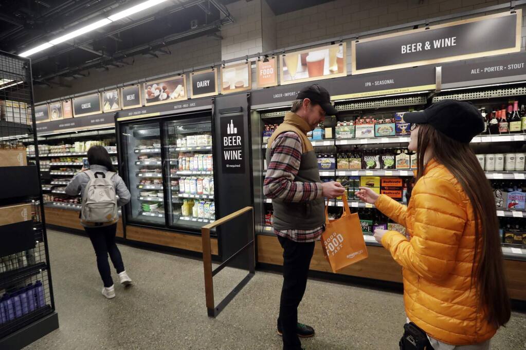 FILE- In this Jan. 22, 2018, file photo, a worker, right, looks at the ID of a shopper at the wine and beer area inside an Amazon Go store in Seattle. Get ready to say good riddance to the checkout line. A year after Amazon opened its first cashier-less store, startups and retailers are racing to get similar technology in other stores throughout the world, letting shoppers buy groceries without waiting in line. (AP Photo/Elaine Thompson, File)