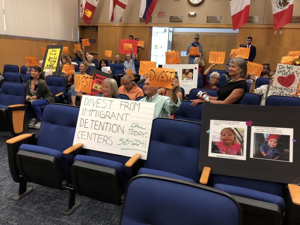 Residents hold up 'Divest' signs Tuesday before the public comment portion of a Board of Sonoma County Supervisors meeting. Residents urged supervisors to divest from U.S.-Mexico border detention facility-connected companies. (Tyler Silvy/The Press Democrat)