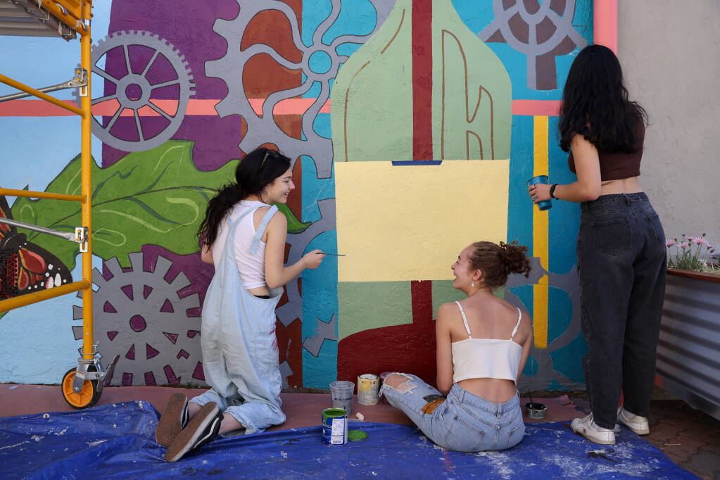 Volunteers Jazmin Zeinalzadehgan, left, Devyn Thierry, and Jessica Simmons paint a mural on Friday, June 24, 2022, on the exterior side wall of Black Oak Coffee Roasters in Healdsburg. (Christopher Chung/The Press Democrat)