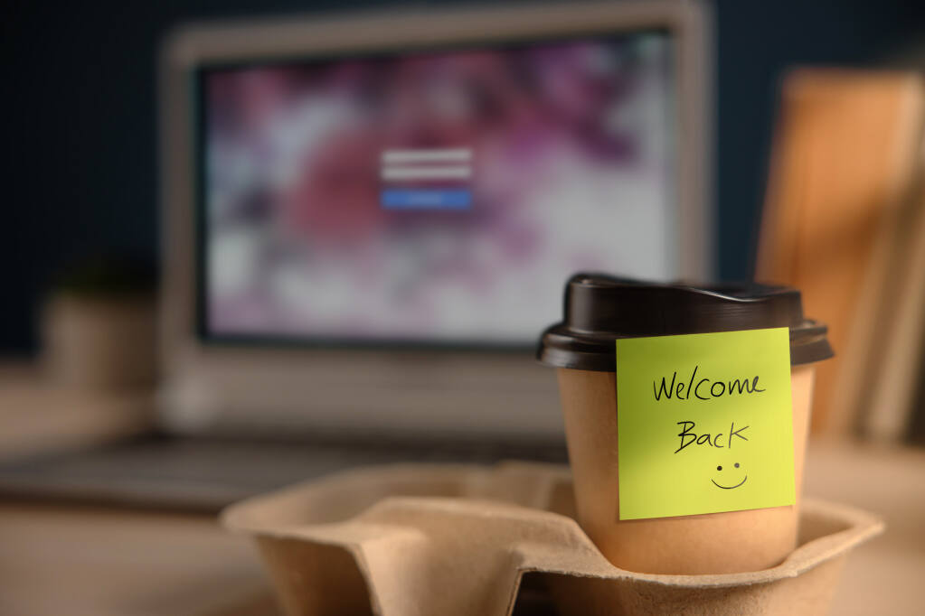 Back to Work concept photo shows a closeup of “welcome back” and a smiley written on a sticky note attached to a coffee cup with black lid in a cardboard carrier, with a computer screen in the background showing an employee log-in screen.