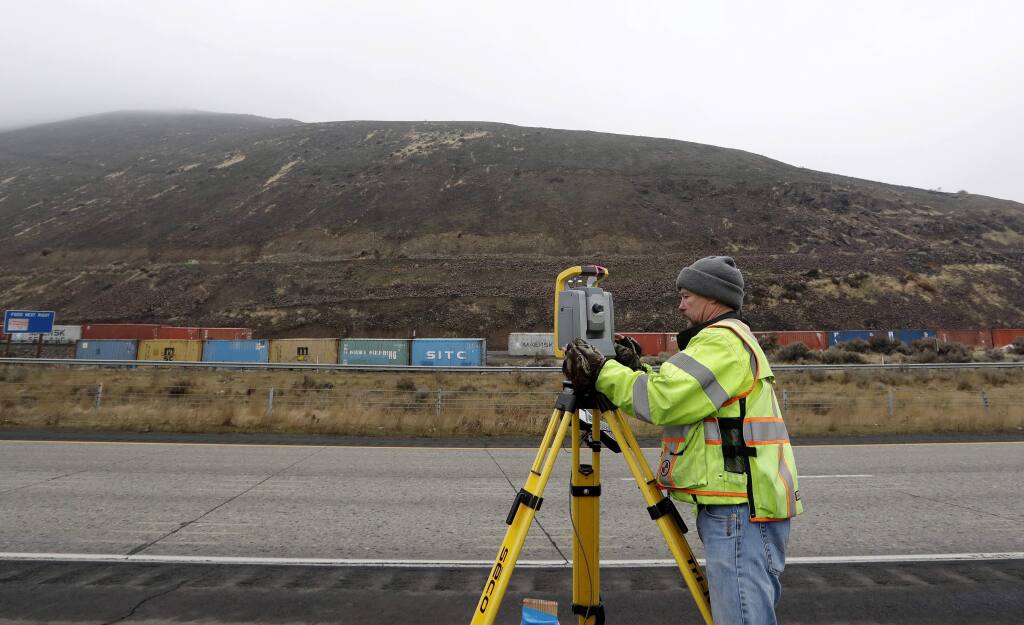 In this Monday, Jan. 8, 2018, photo, James Gleeson, a surveyor with the Washington State Department of Transportation, takes measurements from the shoulder of Interstate 82 to monitor a slow-moving landslide on Rattlesnake Ridge beyond in Union Gap, Wash. Large containers line a road below the ridge, an effort to help block rocks and debris from reaching the highway. The threat has forced evacuations as officials prepare for what they say is inevitable – the collapse of the ridge near the interstate highway that experts say should occur sometime from late January or early February. (AP Photo/Elaine Thompson)