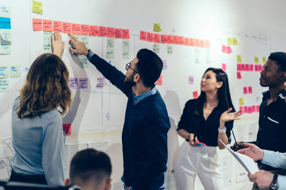 A team of multicultural young male and female professionals in an office point to a project-management wall with colorful paper sticky notes.