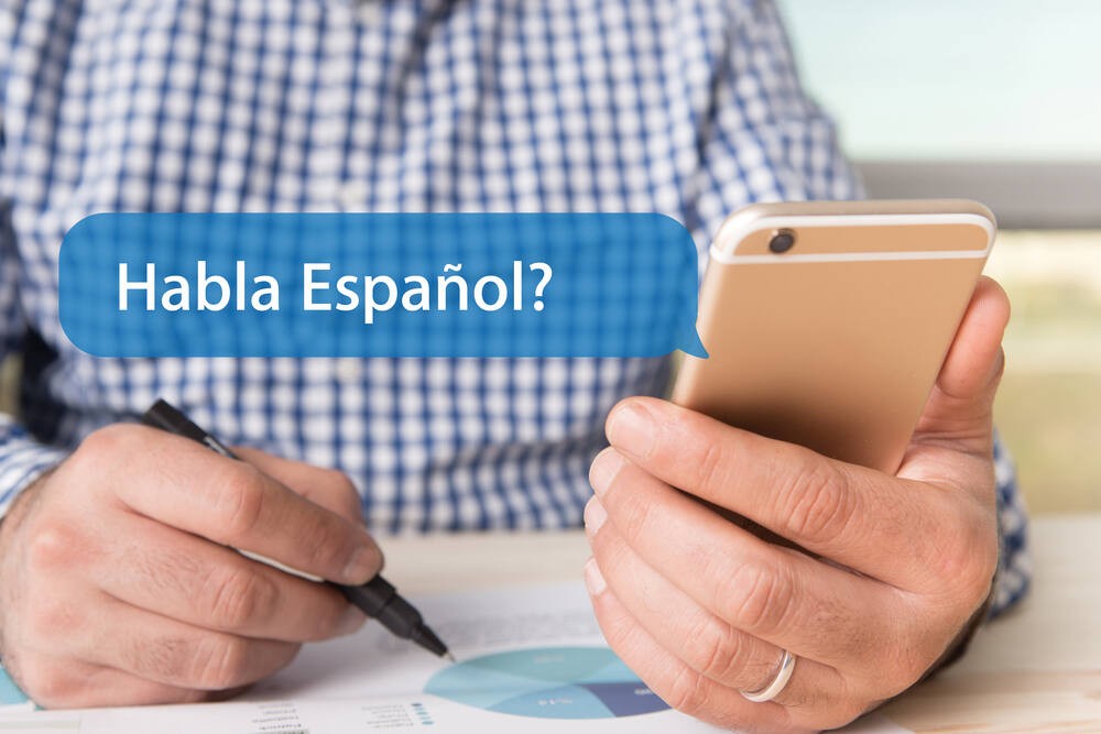 “Habla Espanol?” shown in a chat bubble from a smartphone as a businessperson writes on a business chart. (Garagestock / Shutterstock)