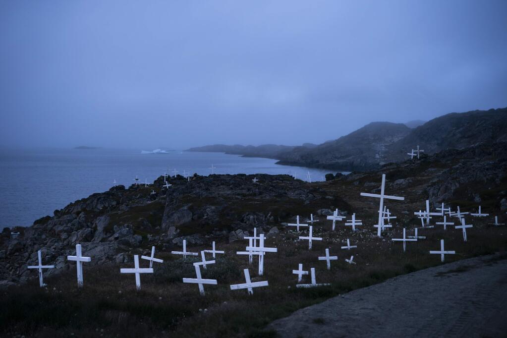 In this Aug. 15, 2019, photo, crosses stand in a cemetery as an iceberg floats in the distance during a foggy morning in Kulusuk, Greenland. Kulusuk's resident Mugu Utuaq says the winter that used to last for as long as 10 months when he was a boy can now be as short as five months. Scientists are hard at work in Greenland, trying to understand the alarmingly rapid melting of the ice. (AP Photo/Felipe Dana)