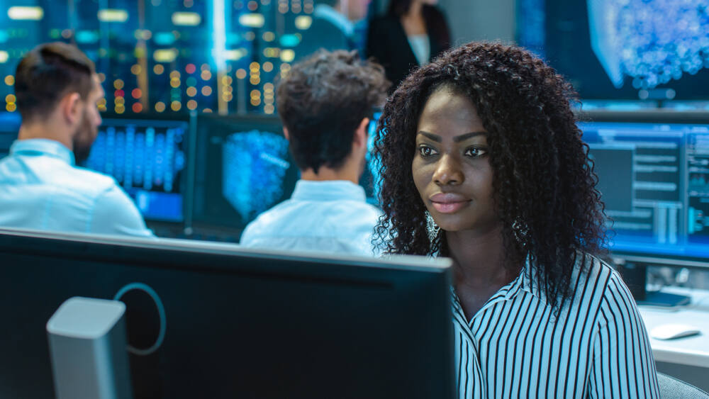 A Black female computer engineer works on a computer in an office with multiple screens and engineers.