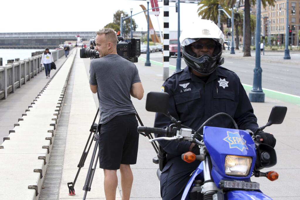 A San Francisco police officer sits on his patrol motorcycle on the Embarcadero by local news cameraman Steve Stifter after a gunman pistol robbed several newscasters of their cameras early Thursday, July 2, 2015 in San Francisco . News crews from two San Francisco Bay Area television stations were robbed at gunpoint and a cameraman was pistol whipped during live broadcasts on Thursday. The robbery and assault occurred at about 6 a.m. along the city's waterfront. Several camera crews were broadcasting live reports for local morning shows about a killing that occurred nearby Wednesday night. (Mike Koozmin/San Francisco Examiner via AP)
