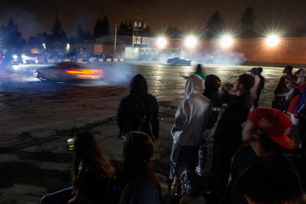 Bystanders watch a pair of drivers turn donuts in their sports cars during a sideshow at the Roseland Village Shopping Center in Santa Rosa, California, on Wednesday, September 16, 2020. (Alvin A.H. Jornada / The Press Democrat)