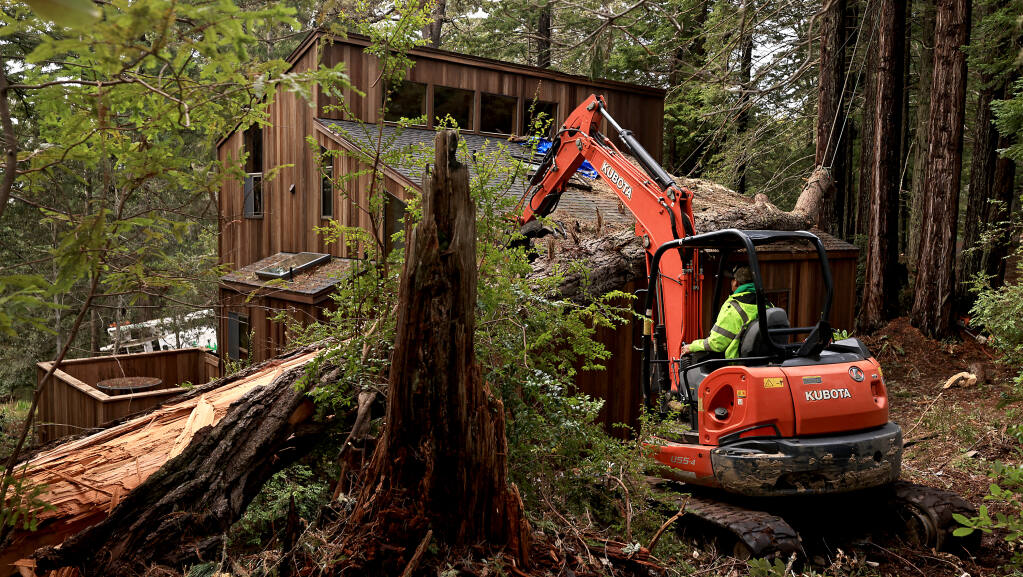 A worker from Gonzalez Tree Service in Gualala, uses a small excavator Thursday, Jan. 12, 2023, to move a large fir tree that fell on a house on Spinnaker Close in the Sea Ranch, in northern Sonoma County, where two people were found dead Wednesday. A generator was found in the home, but no cause of death has been declared. (Kent Porter / The Press Democrat) 2023