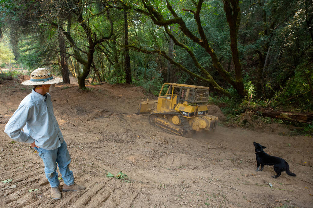 Nick Broderick and his dog, Blue, watch as a firefighting bulldozer rumbles back down a wide fire break, created with bulldozers to defend homes from the Walbridge fire, beside a tributary of Felta Creek in Healdsburg on Wednesday, Aug. 26, 2020. (Alvin A.H. Jornada / The Press Democrat)
