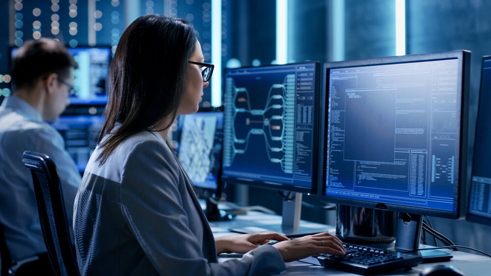 A female information technology professional monitors multiple screens in a blue-lit network or software operations center.
