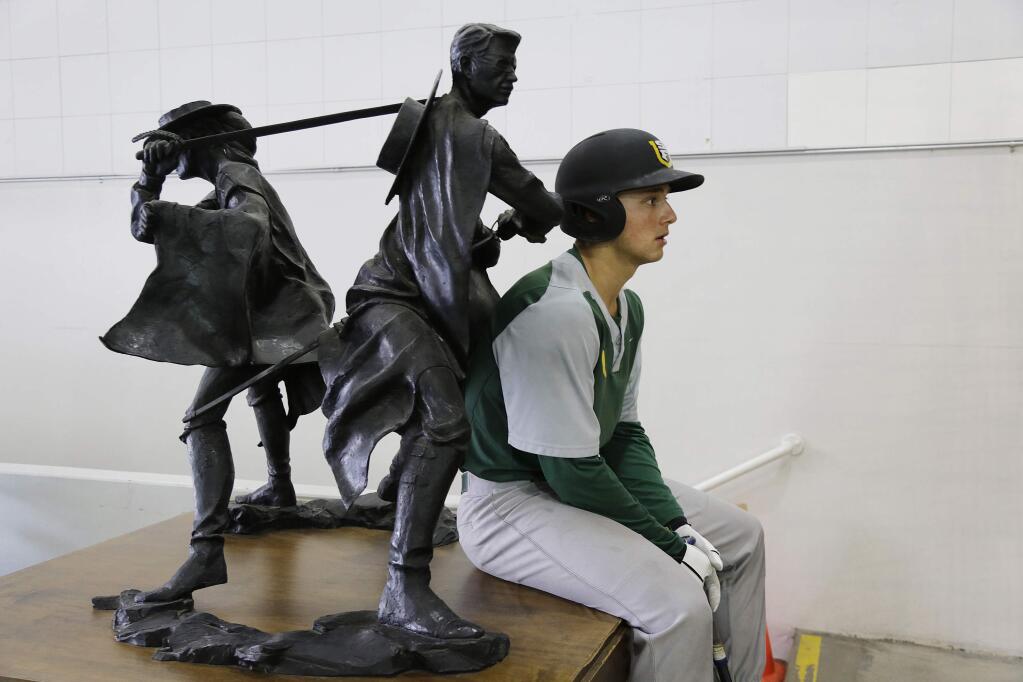In this photo taken Wednesday, March 2, 2016, University of San Francisco infielder Nico Giarratano sits below a statue of Dons while waiting his turn to hit inside inflatable batting cages set up in the school's War Memorial Gym in San Francisco. You can call them the 'VagaDons,' a nickname catching on around campus as they cope with an ever-changing and challenging nomadic lifestyle so far this season while awaiting the finish of its rebuilt campus ballpark, Benedetti Diamond. (AP Photo/Eric Risberg)