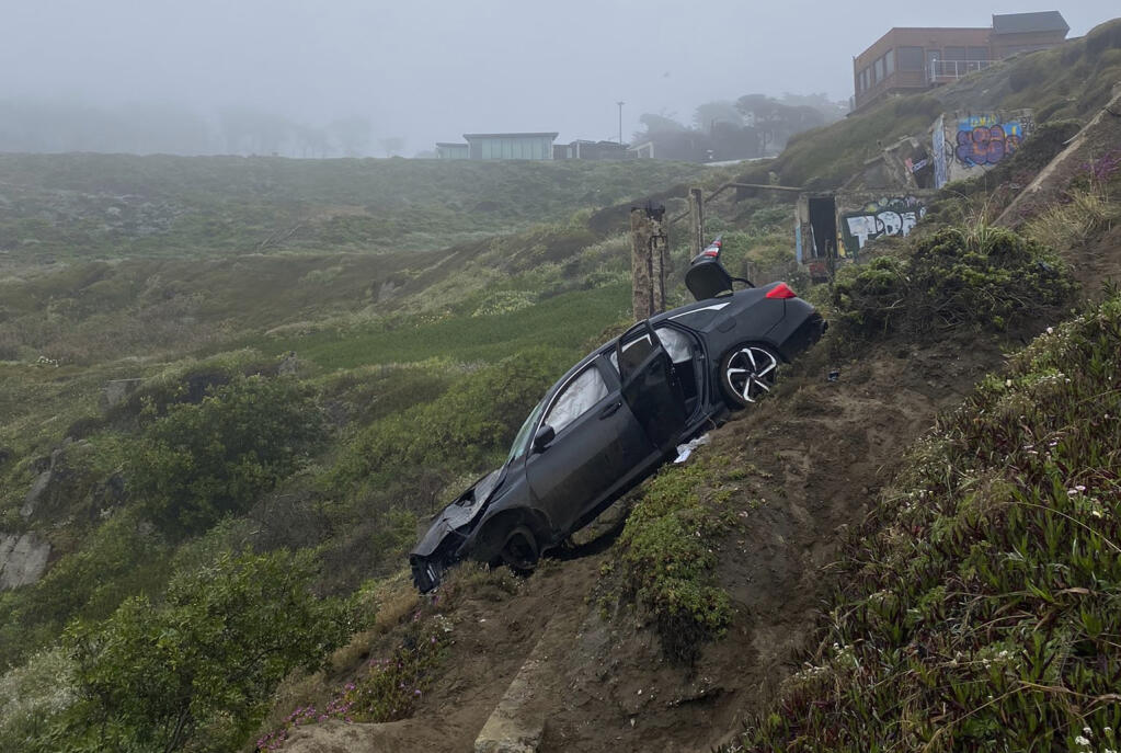 This photo provided by the San Francisco Fire Department shows a dark colored, four-door sedan that went over a cliff near Sutro Baths in San Francisco, Friday, May 6, 2022. Four people were injured when their car plummeted about 120 feet (40 yards) off the cliff in the pre-dawn darkness Friday, authorities said. (San Francisco Fire Department via AP)