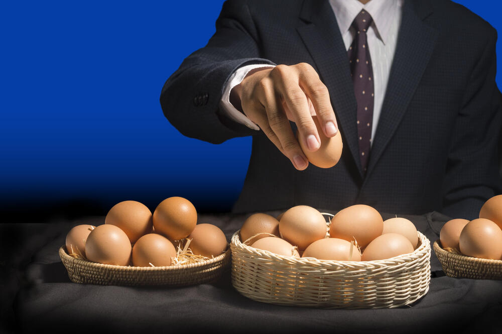 Illustrating the aphorism “Do not put all your eggs in one basket,” a business man holds an egg while moving eggs from many baskets into one.