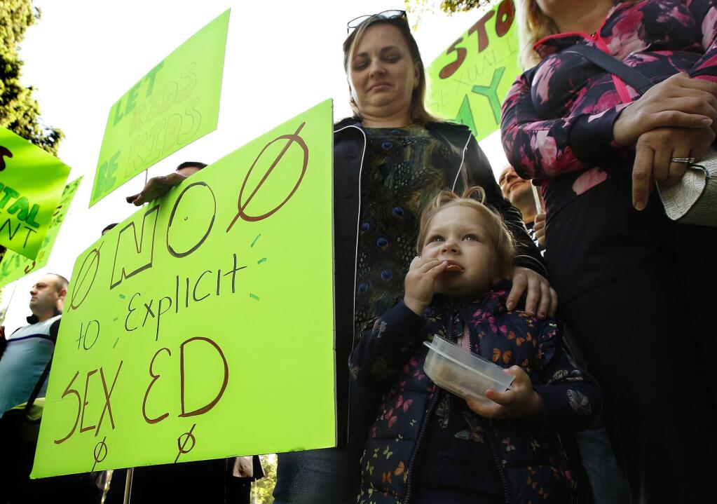 Vicktoryia Seliuzhytskaya, looks down as her daughter Magdlene, 2, eats a snack during a rally to protest proposed changes to sex education guidance for teachers, Wednesday, May 8, 2019, in Sacramento, Calif. The California State Board of Education is set to vote Wednesday on new guidance for teaching sex education in public schools. The guidance is not mandatory but it gives teachers ideas about how to teach a wide range of health topic including speaking to children about gender identity. (AP Photo/Rich Pedroncelli)