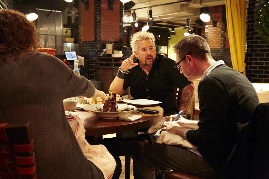 This Dec. 9, 2013 photo shows Food Network star Guy Fieri, center, speaking with AP Food Editor J.M. Hirsch, right, as a plate of General Tso's pork shank, foreground, is displayed at his restaurant, Guy's American Kitchen and Bar, on a tour of premier places to get Super Bowl grub in New York. (Photo by Dan Hallman/Invision/AP)