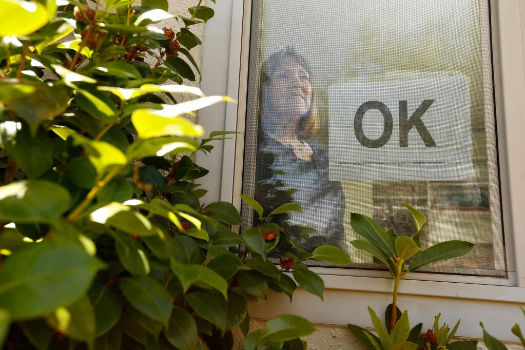 Jill Gordon, chair of the Citizens Organized to Prepare for Emergencies (COPE) committee at Brush Creek Villas, stands in a front window of her home with an 'OK' sign, which she and her neighbors would use during an emergency to show their status, in Santa Rosa, California, on Saturday, June 9, 2018. (Alvin Jornada / The Press Democrat)