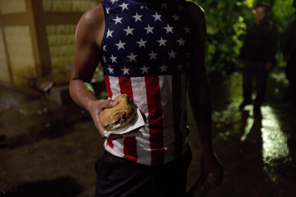 A Honduran migrant wearing a U.S. flag motif shirt, holds a sandwich at an improvised shelter in Chiquimula, Guatemala, Tuesday, Oct. 16, 2018. U.S. President Donald Trump threatened on Tuesday to cut aid to Honduras if it doesn't stop the impromptu caravan of migrants, but it remains unclear if governments in the region can summon the political will to physically halt the determined border-crossers. (AP Photo/Moises Castillo)
