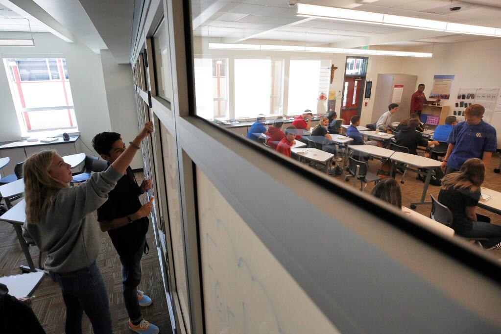 Cardinal Newman junior Reese Lashinski, left, and her classmates use dry-erase markers to write the Catholic cardinal and theological virtues on the movable glass partition wall separating classrooms at Cardinal Newman High School in Santa Rosa, California, on Thursday, August 29, 2019. The new building that houses these classrooms was recently completed to replace one of the campus buildings that burned down during the Tubbs Fire in 2017. (Alvin Jornada / The Press Democrat)