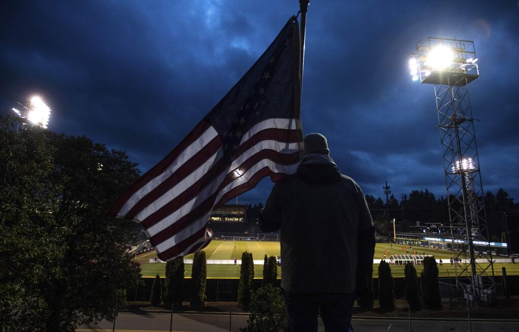 FILE - In this Wednesday, March 11, 2020 file photo, a man holds an U.S. flag as he watches the a United Soccer League match in Tacoma, Wash. In a matter of days, millions of Americans have seen their lives upended by measures to curb the spread of the new coronavirus in mid-March. (Joshua Bessex/The News Tribune via AP)